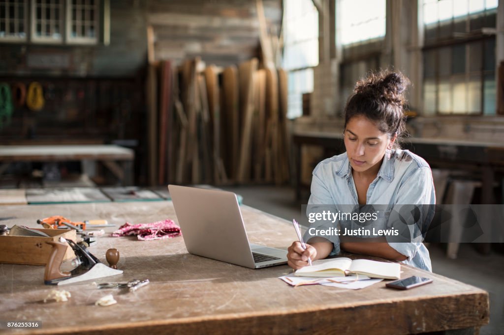 Mixed-race Young Woman Entrepreneur Leans Into Workbench Where She Calculates Solutions to Today's Design Challenges