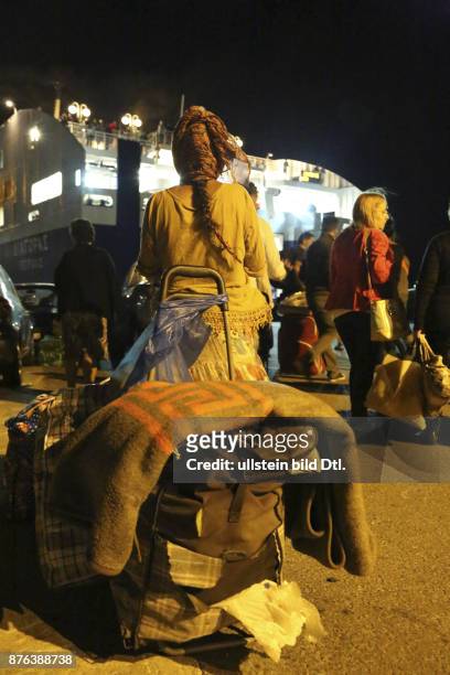 Eine Flüchtlingsfrau wartet mit ihrem Gepäck auf die Fähre nach Piräus. Im Hintergrund die ankommende Fähre. Dunkelheit.