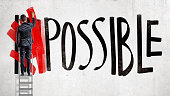 A businessman stands on a stepladder and hides the word Impossible written on the wall using a red paint roller.