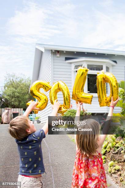 a girl and boy stand in front of house holding gold 'sold' balloons - nz house and driveway stock-fotos und bilder