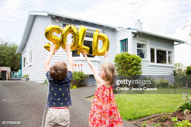 a girl and boy stand in front of house holding gold 'sold' balloons - nz house and driveway stock-fotos und bilder