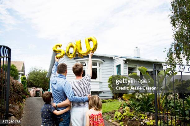 a mum and dad and two kids stand in front of house holding gold 'sold' balloons - nz house and driveway stock-fotos und bilder