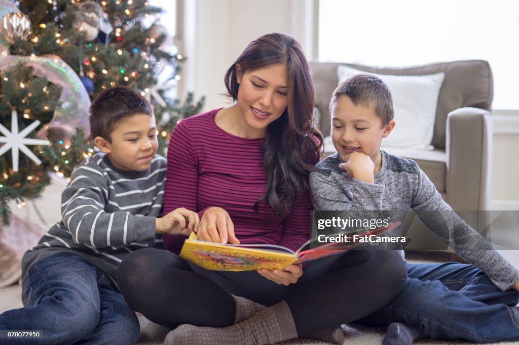 A mom and her two sons read a Christmas book together in front of the decorated tree