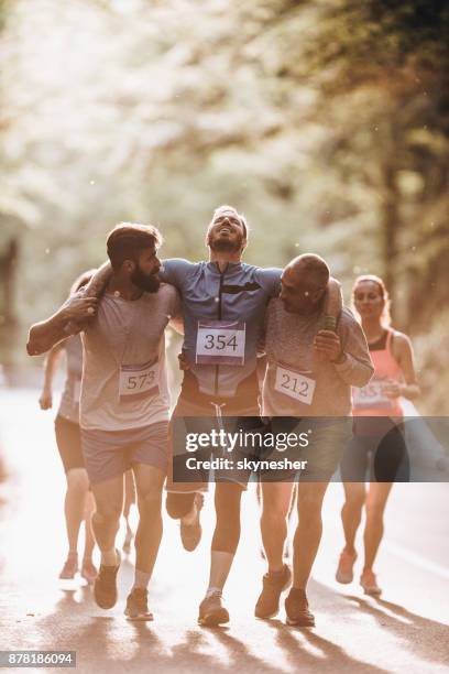 männliche läufer tragen verletzte sportler während der marathon-lauf in der natur. - fairness stock-fotos und bilder