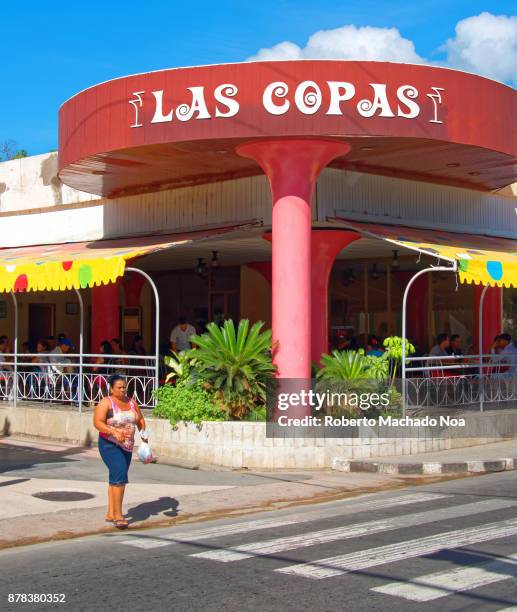 Circular red signage of Las Copas food joint, it has pink pillars as base, a woman crossing road at its front.