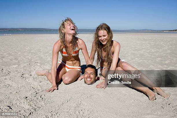 young women burying man in sand laughing - head in sand stock-fotos und bilder