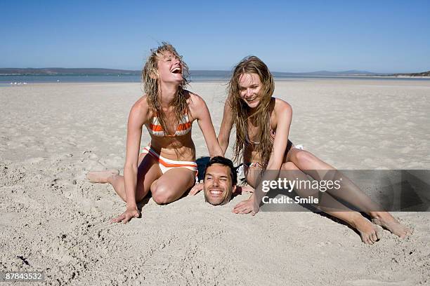 young women burying man in sand laughing - buried in sand stockfoto's en -beelden