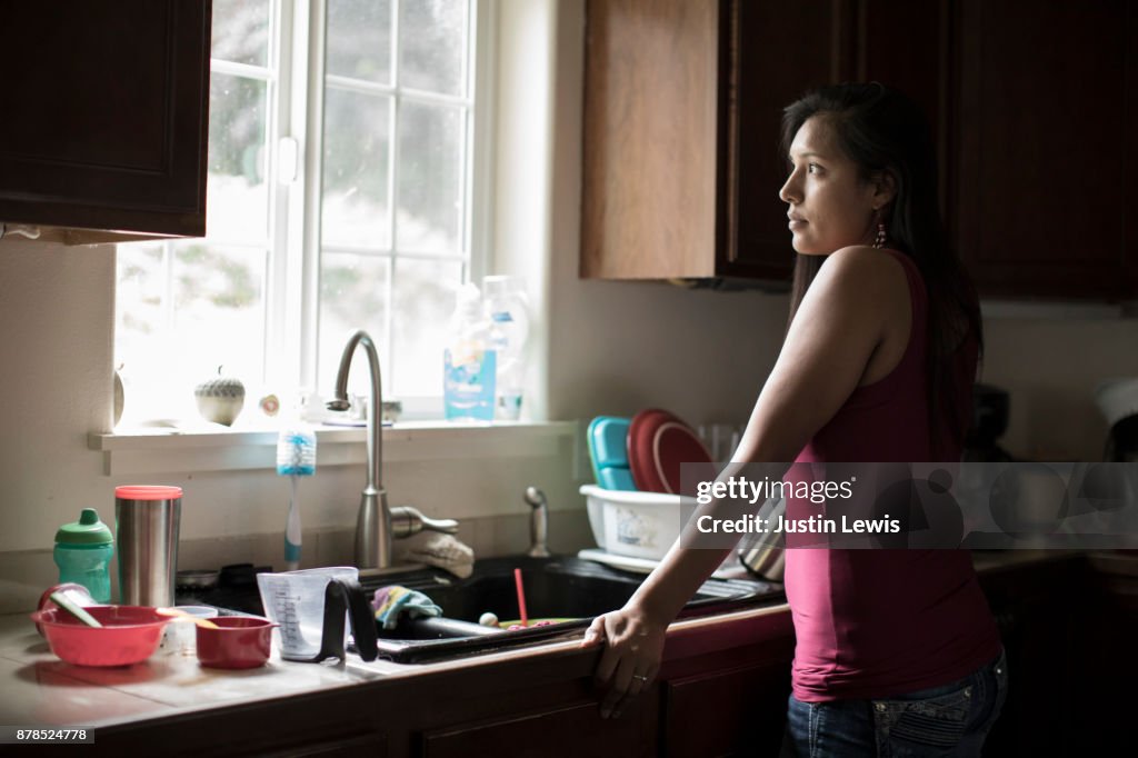 A Young Native American Mother Leans Into the Kitchen Sink, Gazing out the Window to Her Yard