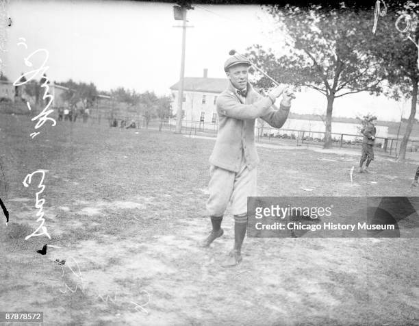 Informal full-length portrait of golfer Charles "Chick" Evans, Jr. Following through after swinging a golf club, standing on an area of patch grass...
