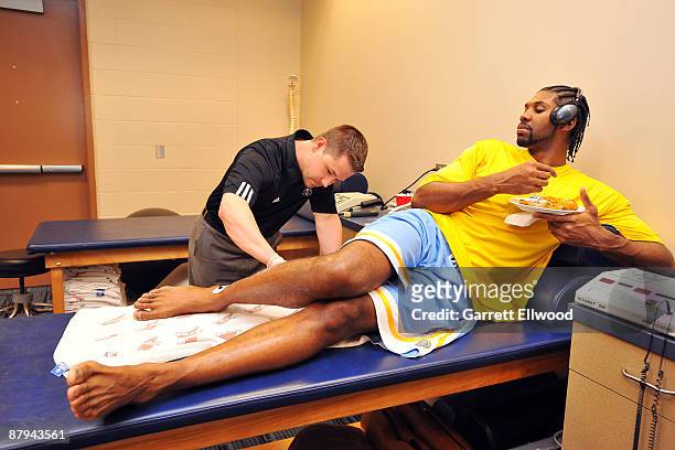 Nene of the Denver Nuggets has a bite to eat while assistant athletic trainer Dan Shimensky goes to work before playing against the Los Angeles...