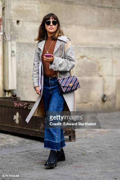 Guest wears sunglasses, a gray trench coat, blue flare jeans, black shoes, a striped bag, outside Koche, during Paris Fashion Week Womenswear...
