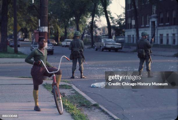 National Guardsmen stand guard as a young boy pauses from riding his bicycle in the west side neighborhood of Lawndale, Chicago, 1968. The guardsmen...