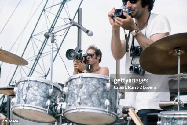 American cinematographer Ted Churchill and photographer Jim Marshall stand near a drum kit and film performers onstage at the Woodstock Music and...