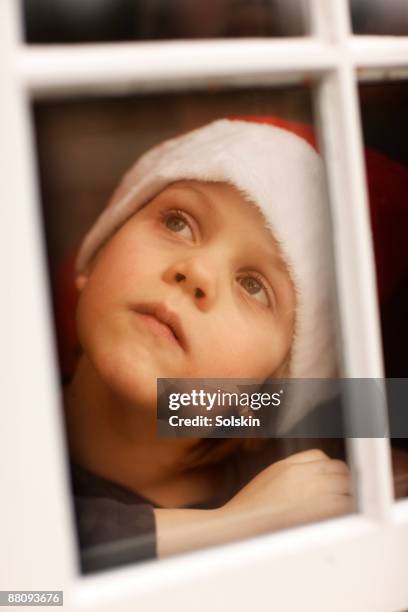 boy with christmas hat, looking out through window - santa leaning stock pictures, royalty-free photos & images