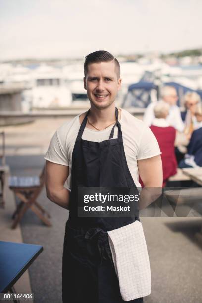 portrait of smiling young waiter wearing apron on restaurant patio - tablier photos et images de collection