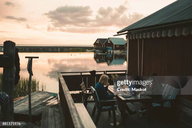 friends using laptop at table in holiday villa by lake against sky - public building stock photos et images de collection