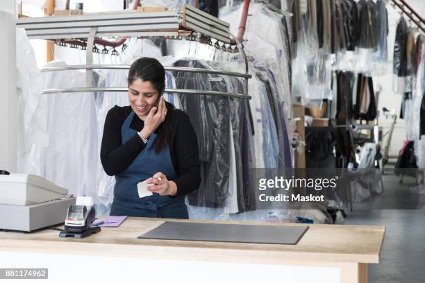 smiling mature female cleaner talking on smart phone while standing at laundromat - stomerij stockfoto's en -beelden