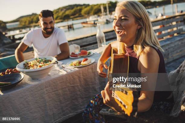 cheerful woman playing guitar while friend sitting at picnic table on sunny day - scandinavia picnic stock pictures, royalty-free photos & images