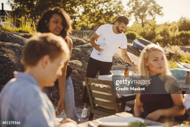 young multi-ethnic friends sitting while man setting table at harbor on sunny day - preparazione al parto foto e immagini stock