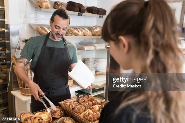 smiling mature male owner serving fresh bread to female customer at bakery - verkäuferin stock-fotos und bilder