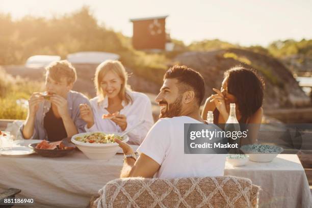 happy friends enjoying lunch at table on jetty - picnic table stock-fotos und bilder