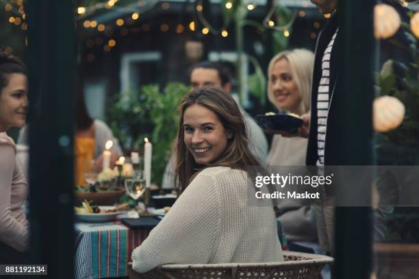 smiling young woman looking back while sitting with friends in glass conservatory in back yard - young women group back stock-fotos und bilder