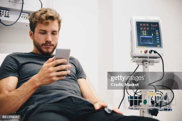 young male patient using smart phone while reclining on bed during medical test in hospital - skane stockfoto's en -beelden