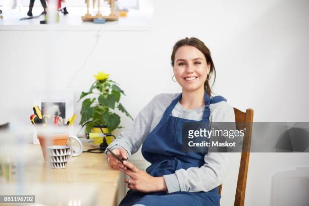 portrait of smiling young female engineer with smart phone sitting at table in workshop - uitvinder stockfoto's en -beelden