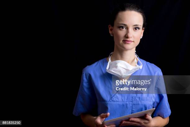 portrait of young female doctor, wearing blue scrubs and a surgical mask and holding a digital tablet, in front of black background. - portrait professional dark background stock pictures, royalty-free photos & images