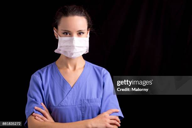 portrait of young female doctor, wearing blue scrubs and a surgical mask, in front of black background. - portrait professional dark background stock pictures, royalty-free photos & images