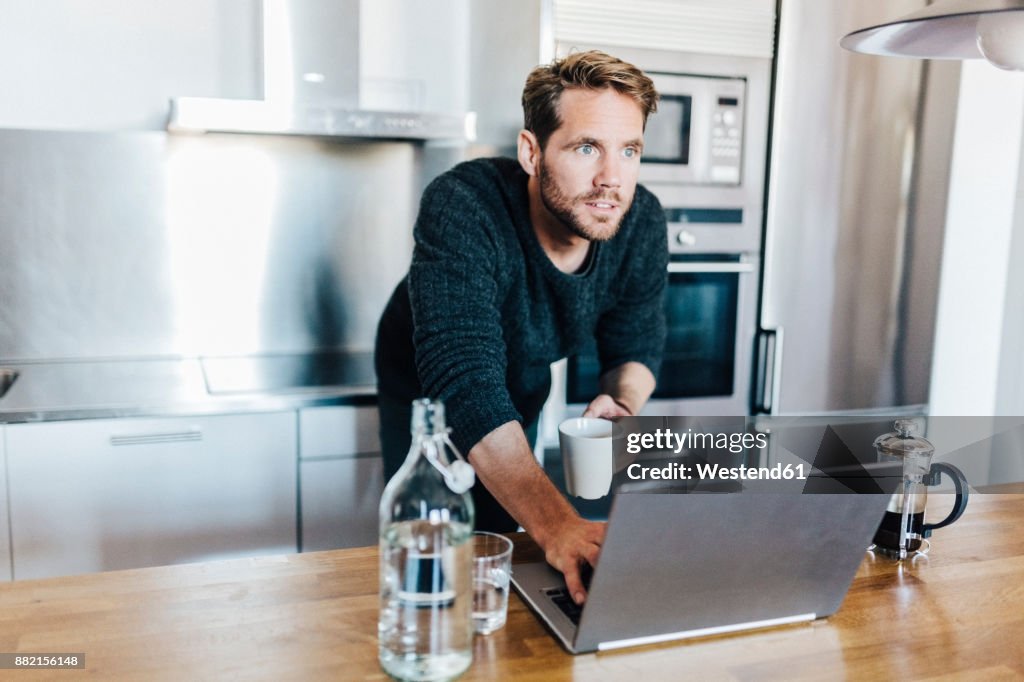 Portrait of starring man with coffee mug and laptop standing in kitchen