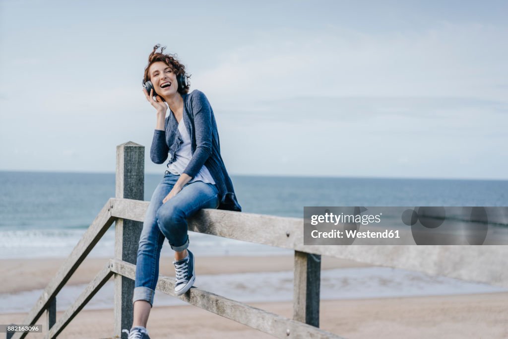Happy woman sitting on railing at the beach listening to music