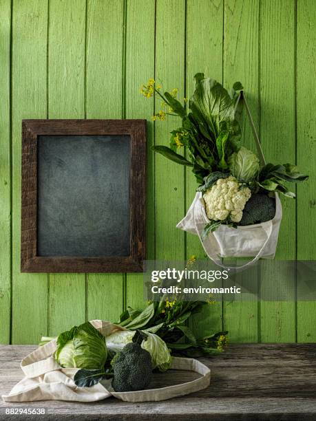 leafy green vegetables hanging in a reusable cotton bag, by the side of a blackboard, on an old green wood board wall background, with another bag of greens on a wood table underneath. - fabric pin board stock pictures, royalty-free photos & images