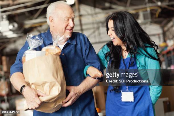 a food bank worker walks arm in arm with a senior patron - hunger food bank stock pictures, royalty-free photos & images