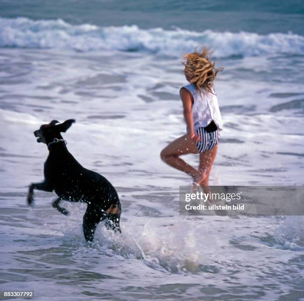 Junges Maedchen laeuft mit Dobermann ausgelassen durch die auslaufende Brandung am Strand