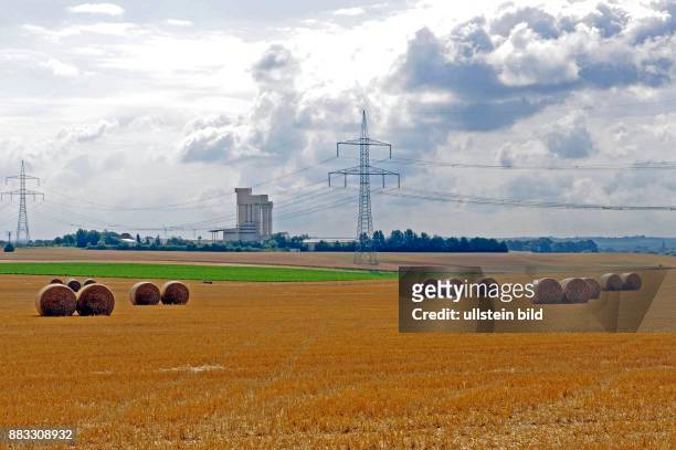 Strohballen liegen auf abgeerntetem Getreidefeld, vor einer Industrieanlage, darueber eine Hochspannungsleitung und Wolkengebirge
