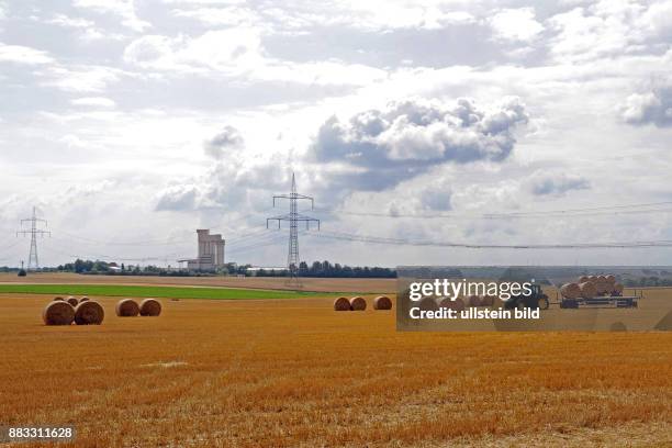 Strohballen liegen auf abgeerntetem Getreidefeld, vor einer Industrieanlage, darueber eine Hochspannungsleitung und Wolkengebirge