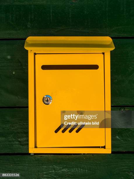 An einer grünen Holzwand ist ein gelber Briefkasten montiert