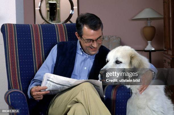 Ein Hundehalter sitzt mit einer Zeitung auf dem Schoß im Sessel und umfaßt einen großen, cremefarbenen Hund, dessen Kopf auf der Armlehne liegt....