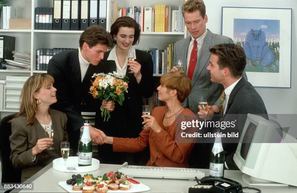 Sektempfang im Büro: Büroangestellte gratulieren einer Kollegin, überreichen einen Blumenstrauß und prosten sich im Rahmen einer kleinen Feier zu