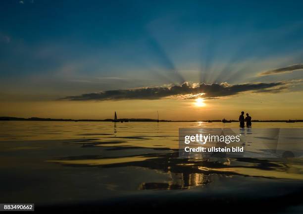 Sonnenstrahlen schimmern durch eine Wolke beim Sonnenuntergang am Chiemsee waehrend zwei Jugendliche im Wasser stehen am 01. Juli 2016 bei Uebersee.