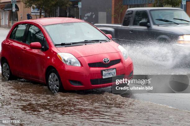 Subcompact car parked next to a flooded sidewalk in Pacific Beach, in the background a Pickup Truck splashing water on the street.