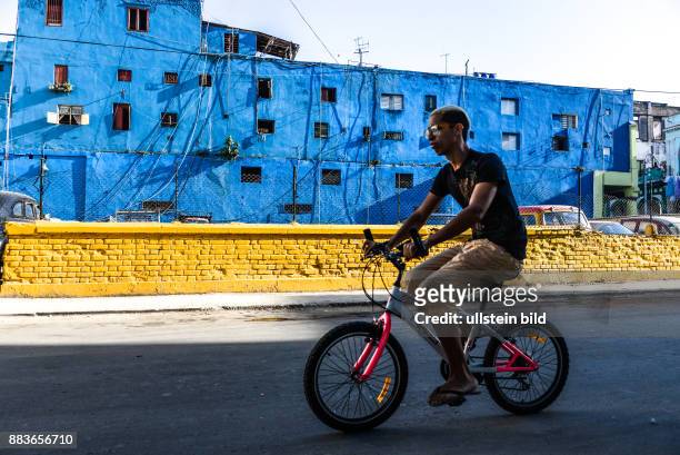 Ein Radfahrer strampelt auf einem Kinderfahrrad durch Havanna. Die blaue Farbe an den Wohnhäusern schützt gegen tropische Hitze