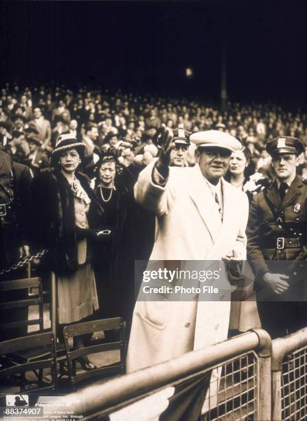 American baseball player George Herman 'Babe' Ruth , dressed in a white overcoat, waves to fans from the front row in an undentified baseball...