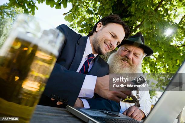 germany, bavaria, upper bavaria, senior bavarian man and young businessman with laptop in beer garden - biergarten stock-fotos und bilder