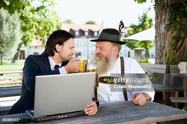 germany, bavaria, upper bavaria, senior bavarian man and young businessman with laptop in beer garden - bierkrug stock-fotos und bilder