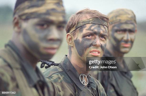 Three US Army soldiers wearing camouflage uniforms and face paint, Vietnam, 1968. Soldier in focus is wearing a peace symbol necklace.