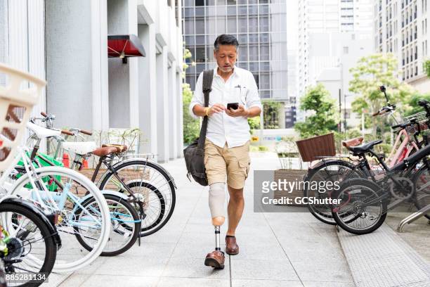 homme japonais avec prothèse jambe marchant le long du terrain de stationnement tout en vérifiant son téléphone - jambes hommes photos et images de collection