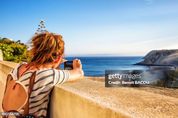 young woman from behind, takes a photo with his smartphone on a low wall from the beach of burgau, algarve region, portugal - burgau portugal stock pictures, royalty-free photos & images