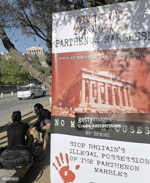 Tourists sit outside the Acropolis hill as a banner reads 'Give back the looted Parthenon marbles, no more excuses, stop Britain's possesion of the...